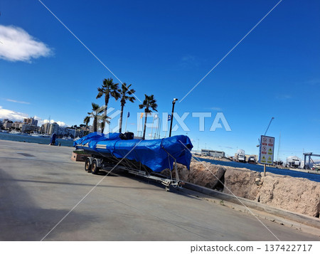 A large motorboat is parked on a trailer at a Mediterranean pier. The boat is covered with a blue tarp. Palm trees line the area, and the sky is clear above the water. 137422717
