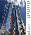 The view shows a contemporary high rise building with a sleek glass facade and orange details. Bright clouds are visible in the sky, and the scene captures mid-day light on the structure. 137422718