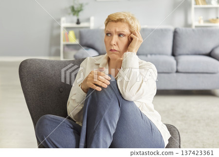 Upset mature worried woman sitting on chair in living room at home with unhappy face expression. Upset mature worried woman sitting on chair in living room at home with unhappy face expression. 137423361