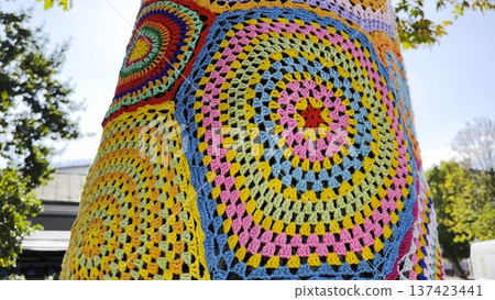 A tree trunk wrapped in colorful knitted cover stands on city street. Handmade yarn bombing installation transforms natural surface into vibrant piece of urban art, blending creativity and texture 137423441