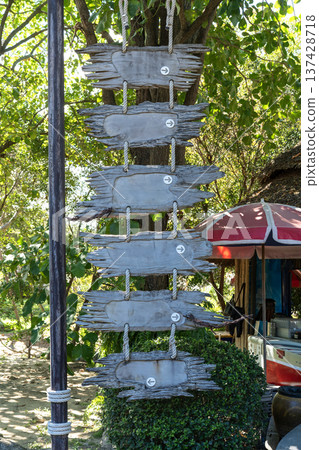 Blank rustic wooden hanging signs with directional arrows on ropes against a lush tropical park background. Vertical row of weathered timber planks for travel navigation or beach resort information. Blank rustic wooden hanging signs with directional arrows on ropes against a lush tropical park background. Vertical row of weathered timber planks for travel navigation or beach resort information. 137428718
