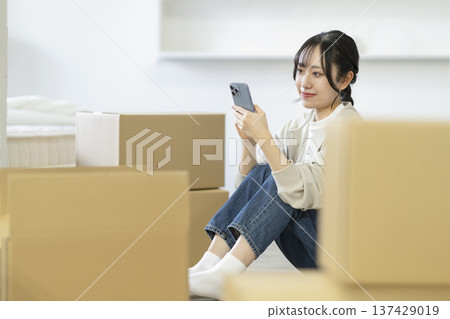 一位年輕女子坐在被紙箱包圍的地方，看著她的智慧型手機。動態影像。 137429019