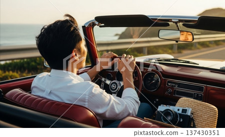 Young Asian man driving classic red convertible car on coastal highway during golden hour sunset with vintage camera 137430351