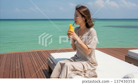 Asian woman drinking orange juice cocktail on luxury resort deck overlooking turquoise ocean water during tropical vacation 137430358