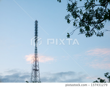Tall, metallic lattice communication tower silhouetted against a dramatic evening sky with surrounding tree branches and leaves, technology and nature conflict. Tall, metallic lattice communication tower silhouetted against a dramatic evening sky with surrounding tree branches and leaves, technology and nature conflict. 137431279