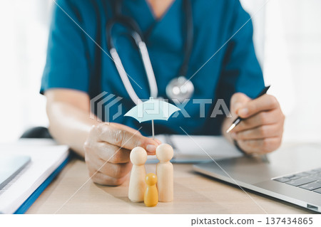 Healthcare worker protects wooden family figures with mini umbrella at clinic desk with stethoscope and laptop, symbolizing health insurance, risk coverage and family care planning today. insurance 137434865