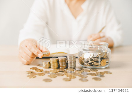 Close-up of hand putting coin into clear glass jar filled with change on bright desk. Minimal home scene for personal savings, budgeting, money management, future goals and security. money management Close-up of hand putting coin into clear glass jar filled with change on bright desk. Minimal home scene for personal savings, budgeting, money management, future goals and security. money management 137434921