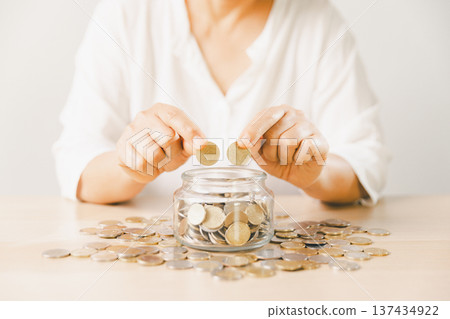 Close-up of hand putting coin into clear glass jar filled with change on bright desk. Minimal home scene for personal savings, budgeting, money management, future goals and security. money management 137434922