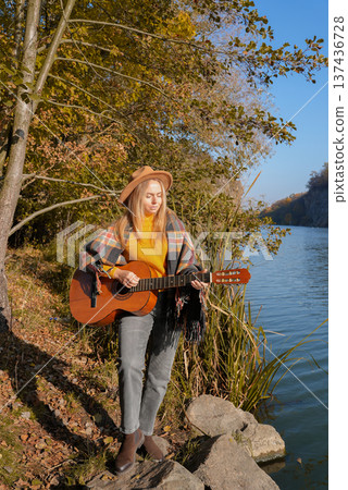 Portrait of smiling caucasian artist plays wooden acoustic guitar in autumnal park. Young songwriter plays string musical instrument hobby outside in nature fall time. Audio music healing Portrait of smiling caucasian artist plays wooden acoustic guitar in autumnal park. Young songwriter plays string musical instrument hobby outside in nature fall time. Audio music healing 137436728