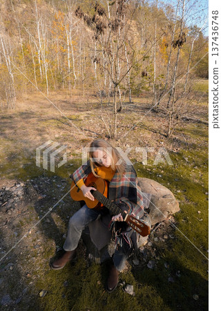 Beautiful blonde girl playing acoustic guitar and singing relaxing enjoying outdoors on sunny autumn day. Hippie woman in boho style learning and practicing playing musical instrument. Fall leaves 137436748