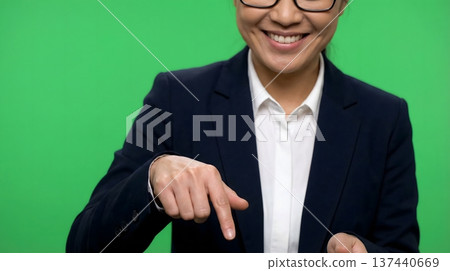 Smiling Asian businesswoman in a suit points down against a green screen background. Ideal for presentations and digital content. Smiling Asian businesswoman in a suit points down against a green screen background. Ideal for presentations and digital content. 137440669