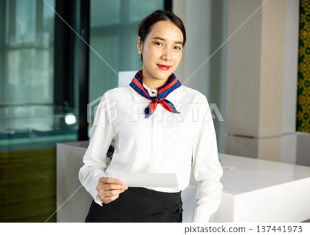Professional airline staff holding boarding pass while standing at check in counter in modern lobby. Friendly hospitality staff providing customer service 137441973