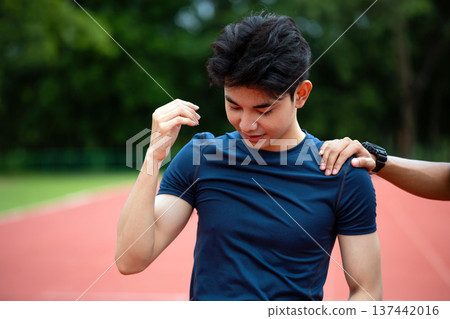 Tired young male runner standing on running track after workout while friend encouraging him. Close up of athlete resting during outdoor training session in stadium track Tired young male runner standing on running track after workout while friend encouraging him. Close up of athlete resting during outdoor training session in stadium track 137442016