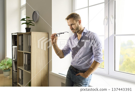 Thoughtful man by large office window, examining glasses and reflecting on choices 137443391