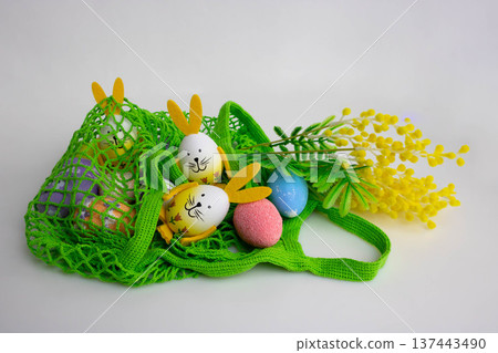 Easter eggs with bunny ears and artificial mimosa branches are lying on the table in a green net. The concept is the Orthodox holiday of Bright Easter 137443490