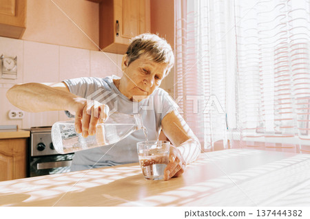 An elderly woman pours clean water into a clear glass while sitting at the table in her cozy kitchen. Disease prevention, maintaining a drinking regimen and maintaining water balance 137444382