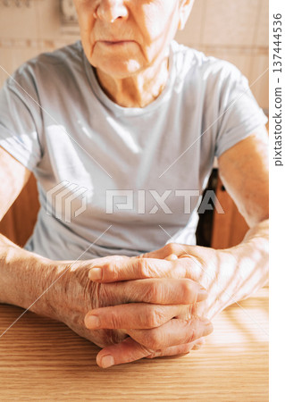 An elderly woman sits at a table in a sunlit room, her hands folded on the table, close-up of her hands 137444536