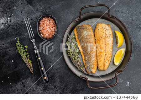 Fried sea bass fillets served on ceramic plate with fork and herbs, overhead restaurant style seafood dish ready to eat with dramatic dark backdrop. 137444560