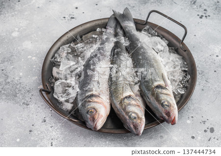 Raw sea bass fish preserved on ice in rustic pan, clean top view composition ideal for fresh seafood advertising, restaurant menu and retail concepts. 137444734
