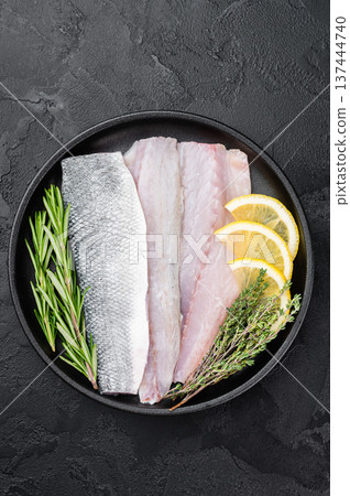 Uncooked European sea bass fillet on black plate with rosemary and thyme, clean culinary layout ideal for seafood menu, cooking recipe or nutrition design. 137444740