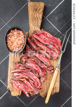 Grilled beef skirt steak slices with coarse salt on wooden cutting board dark textured kitchen background overhead gourmet serving style Grilled beef skirt steak slices with coarse salt on wooden cutting board dark textured kitchen background overhead gourmet serving style 137445016