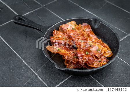 Sizzling bacon slices with rich glaze arranged in black skillet against dark tile background delivering bold gourmet look. 137445132