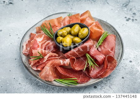 Italian charcuterie platter with bresaola, prosciutto, coppa and green olives arranged with rosemary on a textured gray background. 137445148