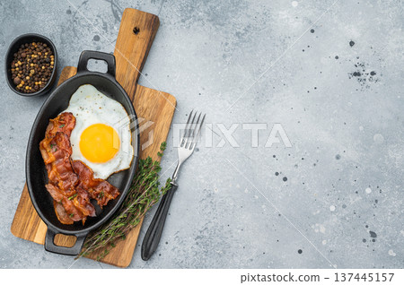 Fried egg with crisp bacon served in black skillet on stone backdrop, flavorful morning meal ideal for keto, brunch and food styling. Fried egg with crisp bacon served in black skillet on stone backdrop, flavorful morning meal ideal for keto, brunch and food styling. 137445157