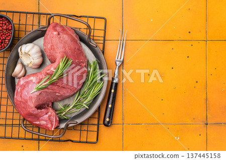 Raw beef tenderloin and roast beef cut with rosemary and garlic in metal pan on yellow background. Fresh meat ingredient with copy space. Raw beef tenderloin and roast beef cut with rosemary and garlic in metal pan on yellow background. Fresh meat ingredient with copy space. 137445158