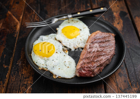 Traditional steak and eggs breakfast with pan fried beef steak and sunny eggs served in cast iron pan, top view on wooden table Traditional steak and eggs breakfast with pan fried beef steak and sunny eggs served in cast iron pan, top view on wooden table 137445199
