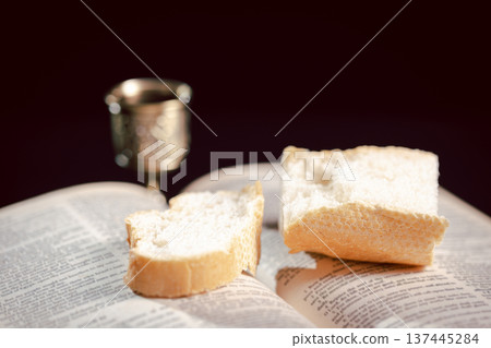 Communion still life with bread on a wooden plate, red wine in a small cup and ornate chalice beside a Holy Bible on dark table, symbolizing Eucharist, worship, faith and remembrance with copy space. 137445284