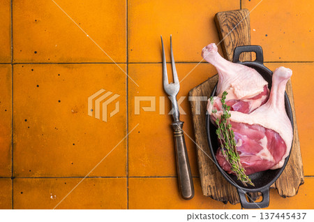 Raw duck legs in a black cast iron skillet, garnished with fresh thyme, resting on a rustic wooden cutting board against an orange tiled background. Raw duck legs in a black cast iron skillet, garnished with fresh thyme, resting on a rustic wooden cutting board against an orange tiled background. 137445437
