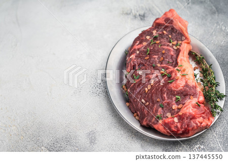 Uncooked strip loin new york beef steak with herbs and coarse salt on a silver plate beside vintage carving fork on soft gray textured background. 137445550