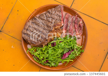 Roasted dry aged striploin new york steak with arugula greens and sliced beef on orange tile background, perfect gourmet dining scene. Roasted dry aged striploin new york steak with arugula greens and sliced beef on orange tile background, perfect gourmet dining scene. 137445601
