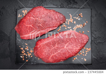 Raw Wagyu chuck eye roll steaks with rosemary and pink salt on dark slate board over textured black background perfect for gourmet butcher stock imagery. Raw Wagyu chuck eye roll steaks with rosemary and pink salt on dark slate board over textured black background perfect for gourmet butcher stock imagery. 137445884