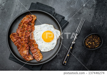 Golden egg yolk with sizzling bacon on matte black dish, dramatic dark surface enhancing texture and shine for top-selling stock images. Golden egg yolk with sizzling bacon on matte black dish, dramatic dark surface enhancing texture and shine for top-selling stock images. 137446274