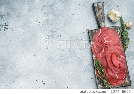 Air dried beef Bresaola charcuterie with marbled red slices, herbs and garlic on a vintage board, top view on a clean stone surface. Air dried beef Bresaola charcuterie with marbled red slices, herbs and garlic on a vintage board, top view on a clean stone surface. 137446665