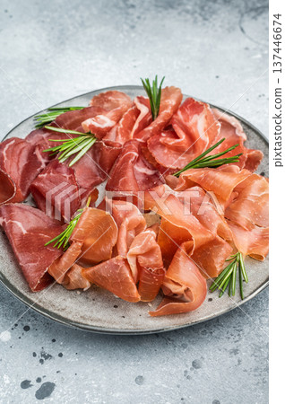 Italian charcuterie platter with bresaola, prosciutto, coppa and green olives arranged with rosemary on a textured gray background. 137446674