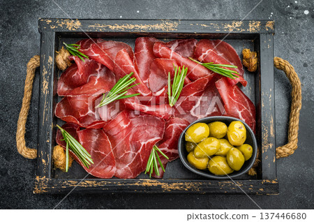 Italian bresaola with rosemary and green olives served in a vintage tray on a dark textured background, shot overhead for gourmet styling. Italian bresaola with rosemary and green olives served in a vintage tray on a dark textured background, shot overhead for gourmet styling. 137446680