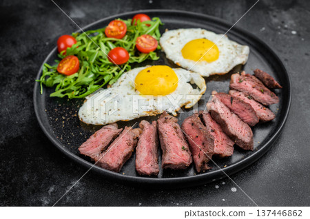 Classic steak and eggs breakfast featuring beef steak slices, sunny side eggs and fresh greens served on black plate over concrete background 137446862