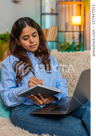 Indian woman studying online with laptop and writing notes in notebook at home, focused expression 137447053