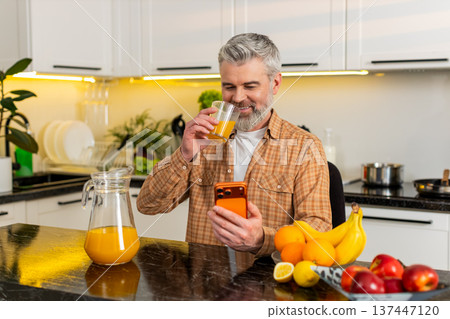 Middle-aged man pours juice drinks from glass checks smartphone news for healthy eating in kitchen Middle-aged man pours juice drinks from glass checks smartphone news for healthy eating in kitchen 137447120