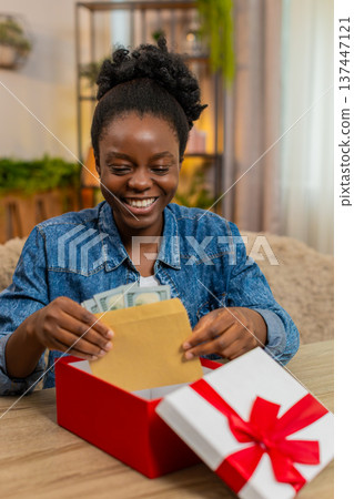 African American young woman opens birthday gift box with envelope of cash dollars sitting at home African American young woman opens birthday gift box with envelope of cash dollars sitting at home 137447121