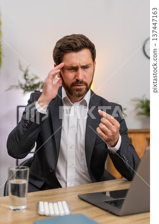 Middle-aged businessman rubs temples swallows pill drinks water resumes typing fast on laptop text 137447163