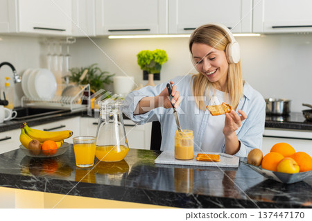 Young woman spreading peanut butter on toast while listening to music with headphones in own kitchen Young woman spreading peanut butter on toast while listening to music with headphones in own kitchen 137447170