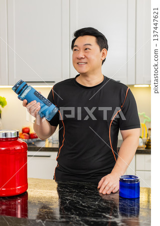 Asian young man shaking shaker bottle and drinking energy supplement drink in home kitchen post gym 137447621