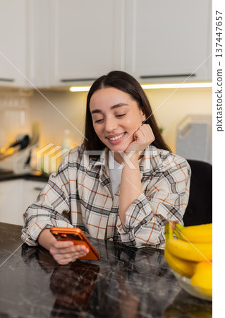 Young woman chats on smartphone at kitchen table scrolling news and buy online alone indoors at home 137447657