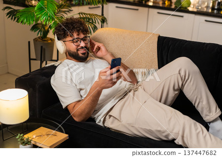 Arabian man wearing wireless headphones listens music texts on smartphone at night indoors relaxed Arabian man wearing wireless headphones listens music texts on smartphone at night indoors relaxed 137447682