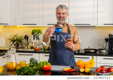 Middle-aged man uses smartphone with credit card in kitchen buying healthy groceries for diet plan Middle-aged man uses smartphone with credit card in kitchen buying healthy groceries for diet plan 137447723