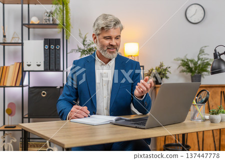 Middle-aged businessman hosts online webinar with earphones via laptop answering audience questions Middle-aged businessman hosts online webinar with earphones via laptop answering audience questions 137447778